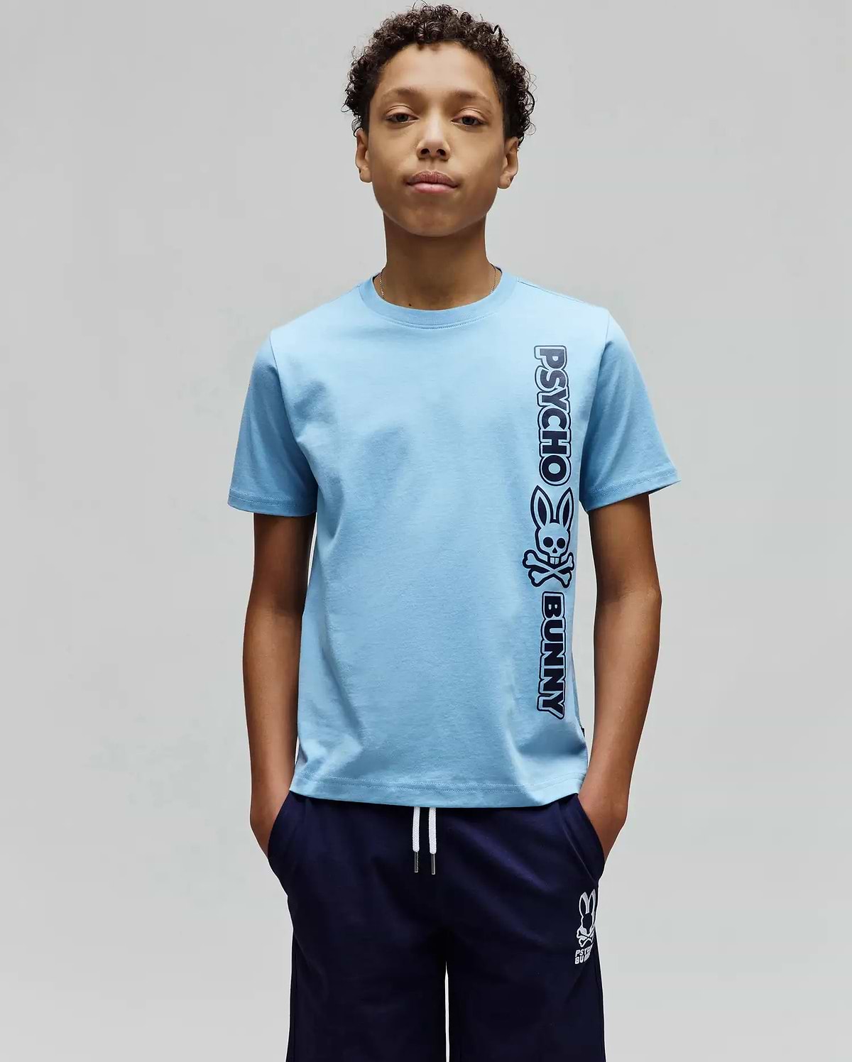 Kids Toby Wordmark Tee, light blue classic fit jersey knit t-shirt with a large vertical dark blue Psycho Bunny wordmark and skull logo graphic on the right side.
