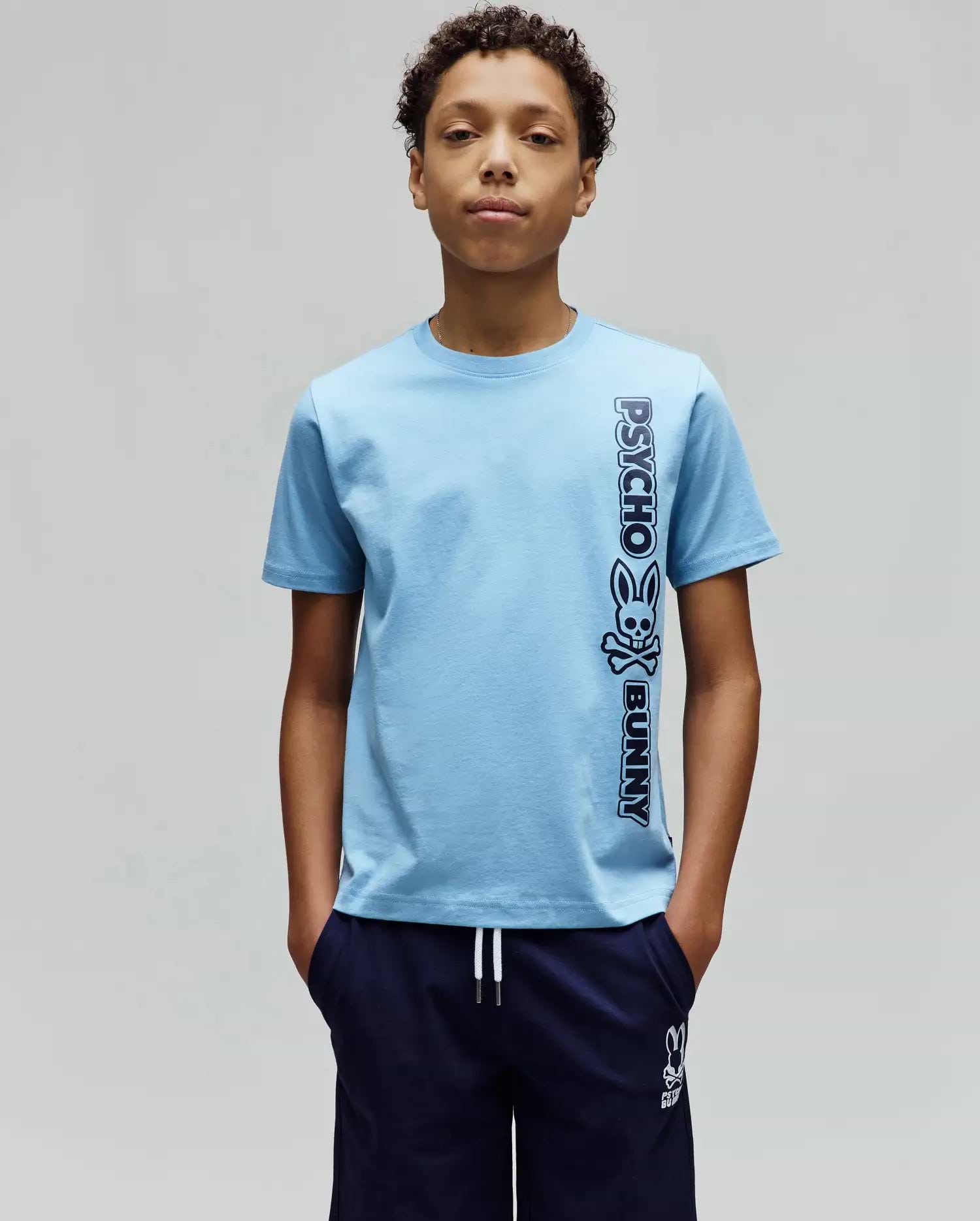 Kids Toby Wordmark Tee, light blue classic fit jersey knit t-shirt with a large vertical dark blue Psycho Bunny wordmark and skull logo graphic on the right side.