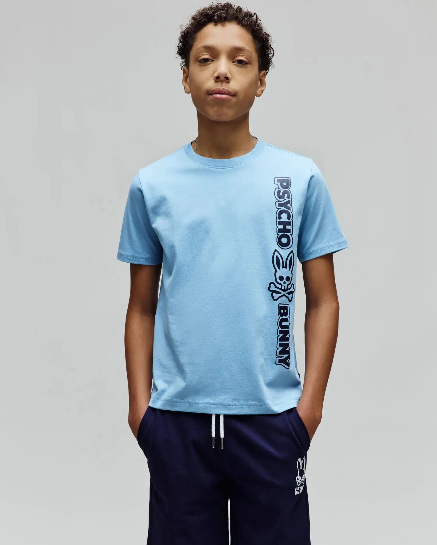 Kids Toby Wordmark Tee, light blue classic fit jersey knit t-shirt with a large vertical dark blue Psycho Bunny wordmark and skull logo graphic on the right side.