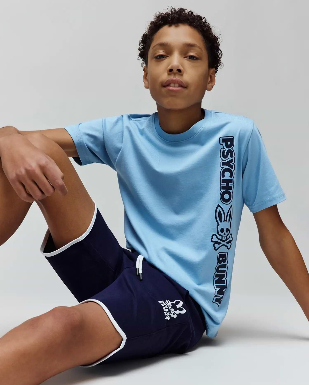 Kids Toby Wordmark Tee in light blue jersey with a large vertical black Psycho Bunny wordmark and skull graphic, paired with navy blue shorts featuring white piping and a small white bunny skull logo.