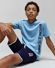 Kids Toby Wordmark Tee in light blue jersey with a large vertical black Psycho Bunny wordmark and skull graphic, paired with navy blue shorts featuring white piping and a small white bunny skull logo.