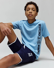 Kids Toby Wordmark Tee in light blue jersey with a large vertical black Psycho Bunny wordmark and skull graphic, paired with navy blue shorts featuring white piping and a small white bunny skull logo.