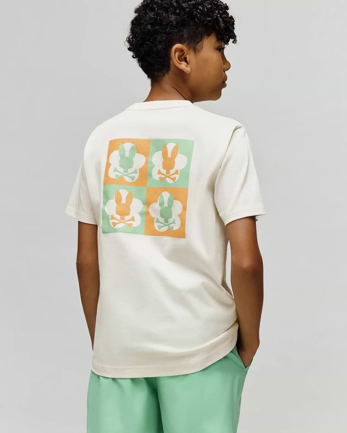 Kids Aiden Back Graphic Tee, a cream classic fit jersey t-shirt with a large back graphic featuring a checkerboard of orange and mint green Psycho Bunny skull logos.