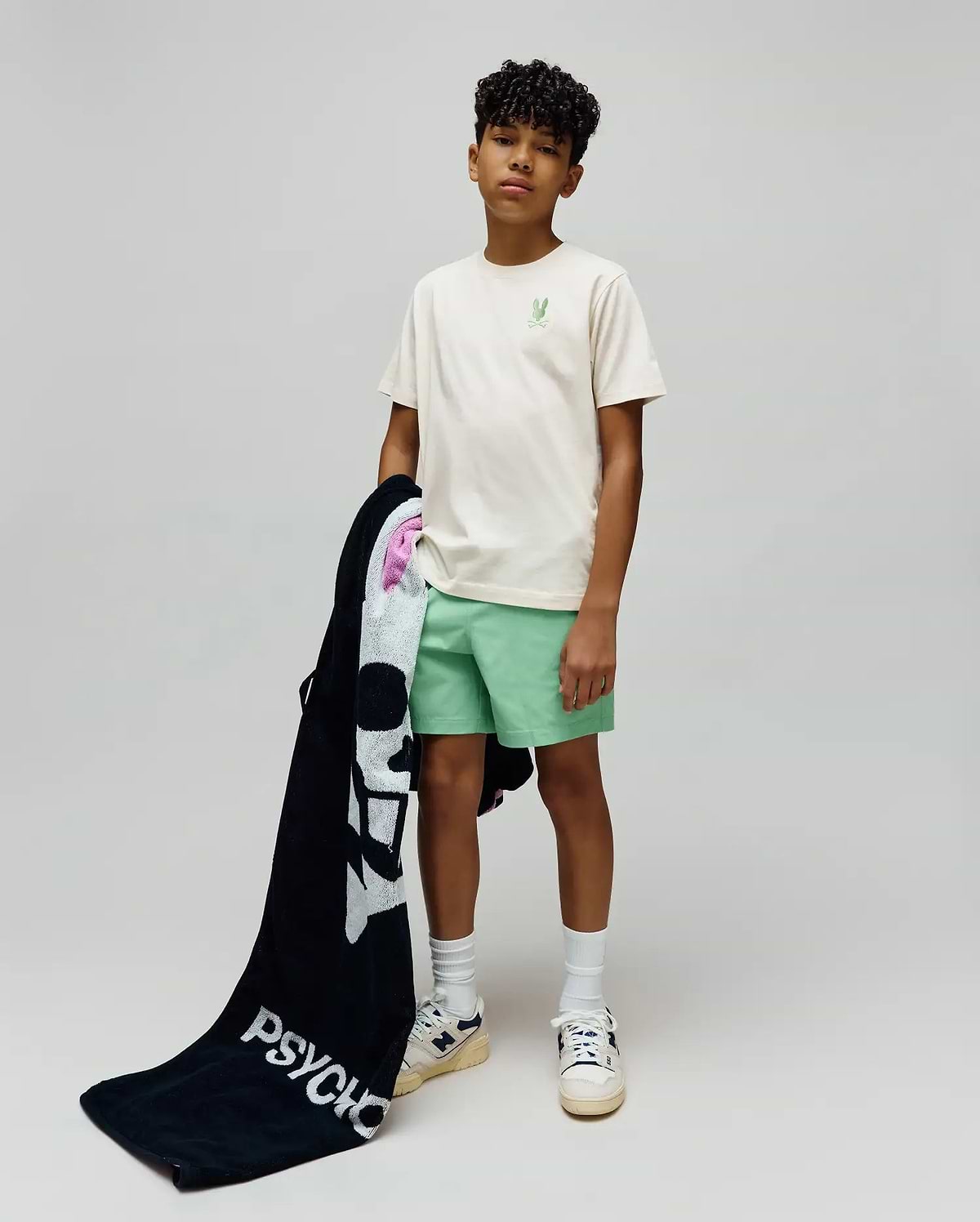 Kids Aiden Back Graphic Tee in ecru jersey with a small embroidered light green Psycho Bunny skull on the chest, paired with mint green shorts and a black terry towel featuring a large white Psycho Bunny skull and PSYCHO text.