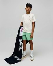 Kids Aiden Back Graphic Tee in ecru jersey with a small embroidered light green Psycho Bunny skull on the chest, paired with mint green shorts and a black terry towel featuring a large white Psycho Bunny skull and PSYCHO text.