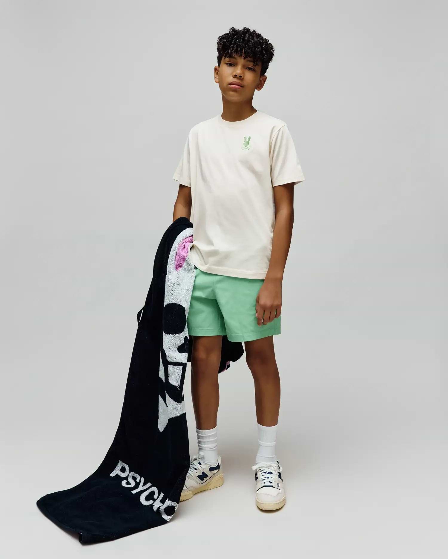 Kids Aiden Back Graphic Tee in ecru jersey with a small embroidered light green Psycho Bunny skull on the chest, paired with mint green shorts and a black terry towel featuring a large white Psycho Bunny skull and PSYCHO text.