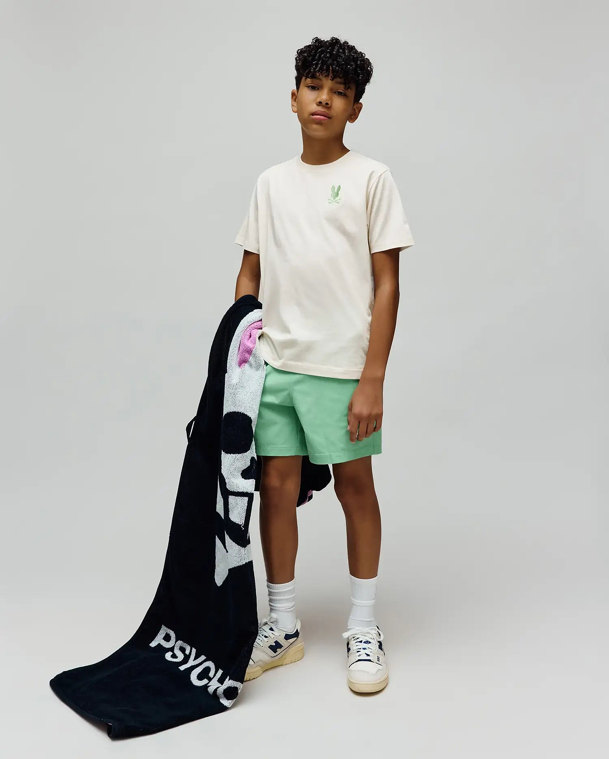 Kids Aiden Back Graphic Tee in ecru jersey with a small embroidered light green Psycho Bunny skull on the chest, paired with mint green shorts and a black terry towel featuring a large white Psycho Bunny skull and PSYCHO text.