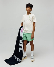 Kids Aiden Back Graphic Tee in ecru jersey with a small embroidered light green Psycho Bunny skull on the chest, paired with mint green shorts and a black terry towel featuring a large white Psycho Bunny skull and PSYCHO text.