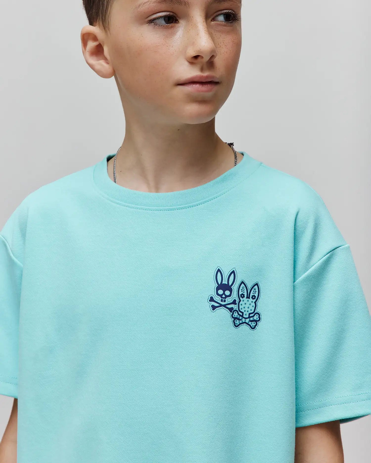 Kids Luke Back Graphic Tee in aqua, featuring a small, embroidered dark blue Psycho Bunny skull logo with two distinct bunnies and crossbones on the left chest, in a relaxed jersey knit.
