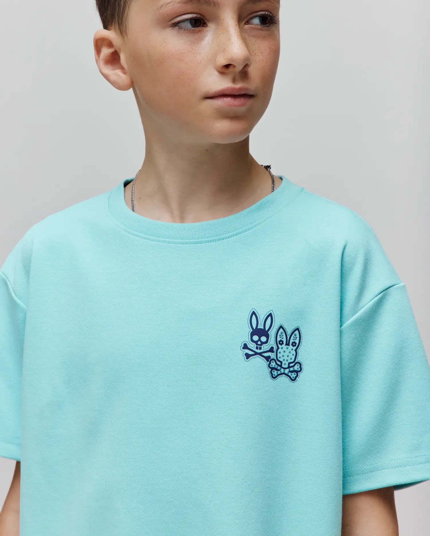 Kids Luke Back Graphic Tee in aqua, featuring a small, embroidered dark blue Psycho Bunny skull logo with two distinct bunnies and crossbones on the left chest, in a relaxed jersey knit.