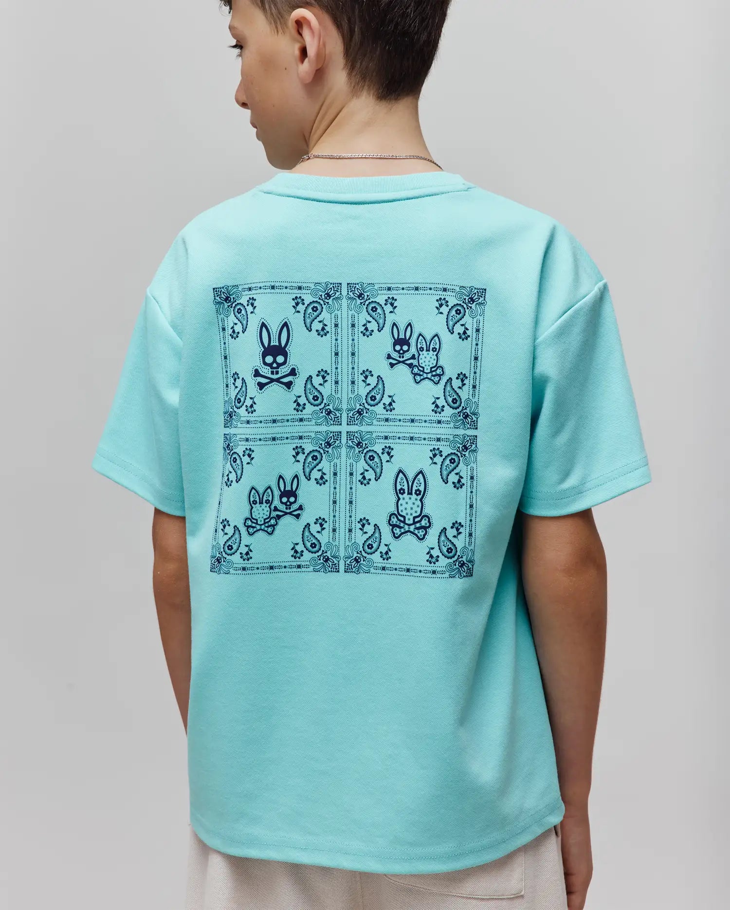 Kids Luke Back Graphic Tee in aqua, featuring a relaxed fit and smooth jersey knit. A large dark blue back graphic displays four squares with bandana borders, paisley patterns, and the iconic Psycho Bunny skull logo.