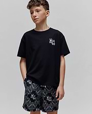 Kids Luke Back Graphic Tee: Black jersey t-shirt with a small white embroidered Psycho Bunny skull logo on the chest. Black relaxed-fit shorts feature an all-over white bandana print with bunny skull motifs.