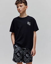 Kids Luke Back Graphic Tee: Black jersey t-shirt with a small white embroidered Psycho Bunny skull logo on the chest. Black relaxed-fit shorts feature an all-over white bandana print with bunny skull motifs.