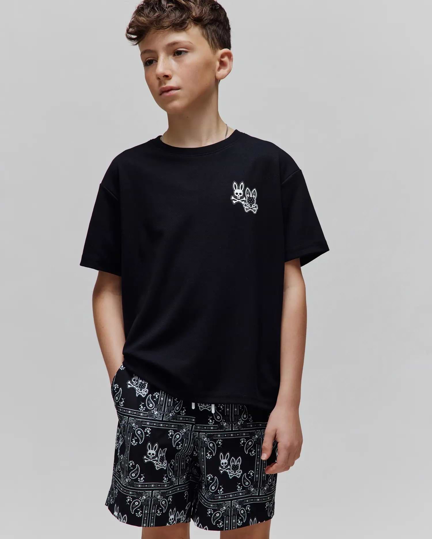 Kids Luke Back Graphic Tee: Black jersey t-shirt with a small white embroidered Psycho Bunny skull logo on the chest. Black relaxed-fit shorts feature an all-over white bandana print with bunny skull motifs.