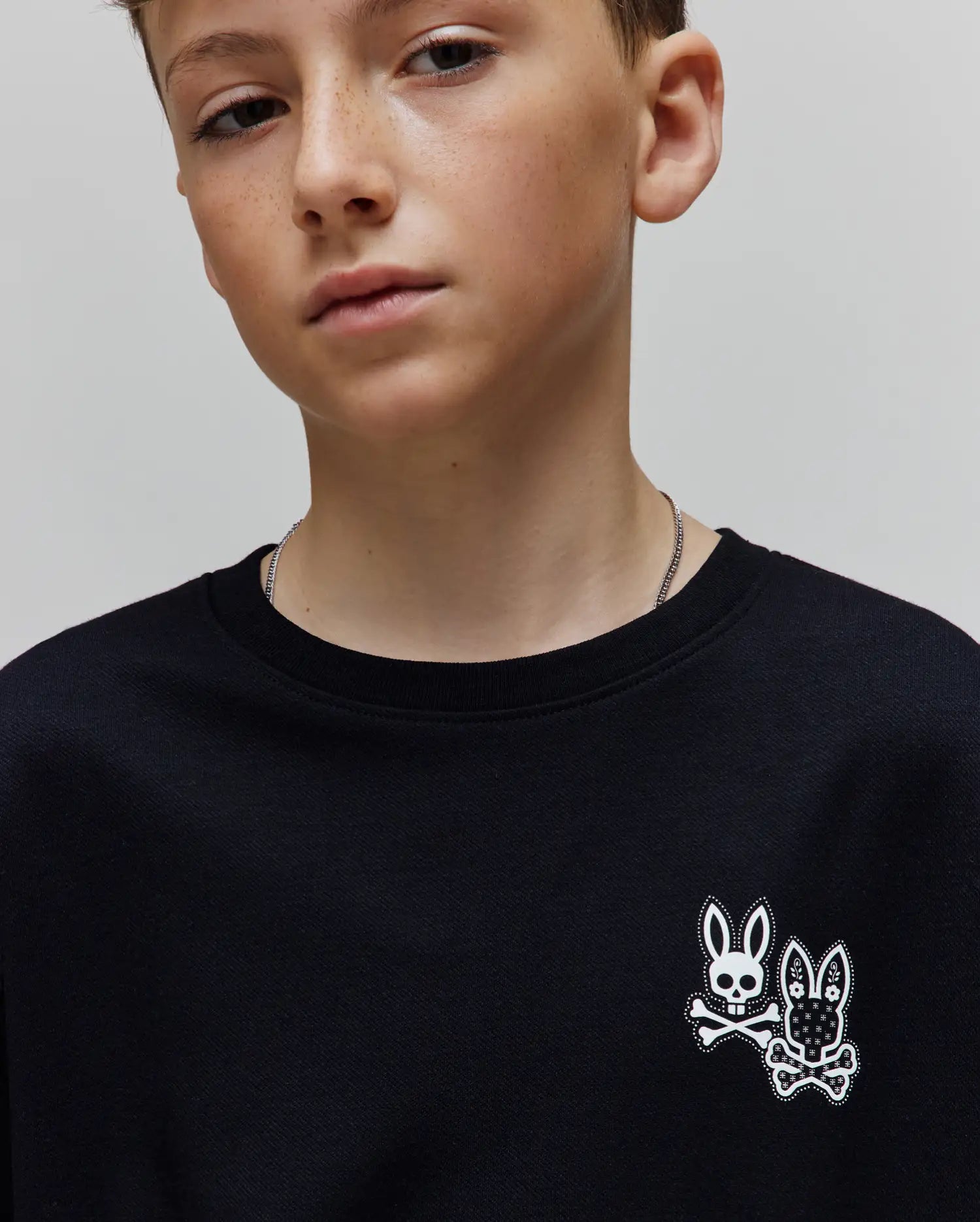 Kids Luke Back Graphic Tee in black jersey knit, featuring a small white double Psycho Bunny skull and crossbones graphic on the front left chest, one classic and one floral patterned, in a classic fit.