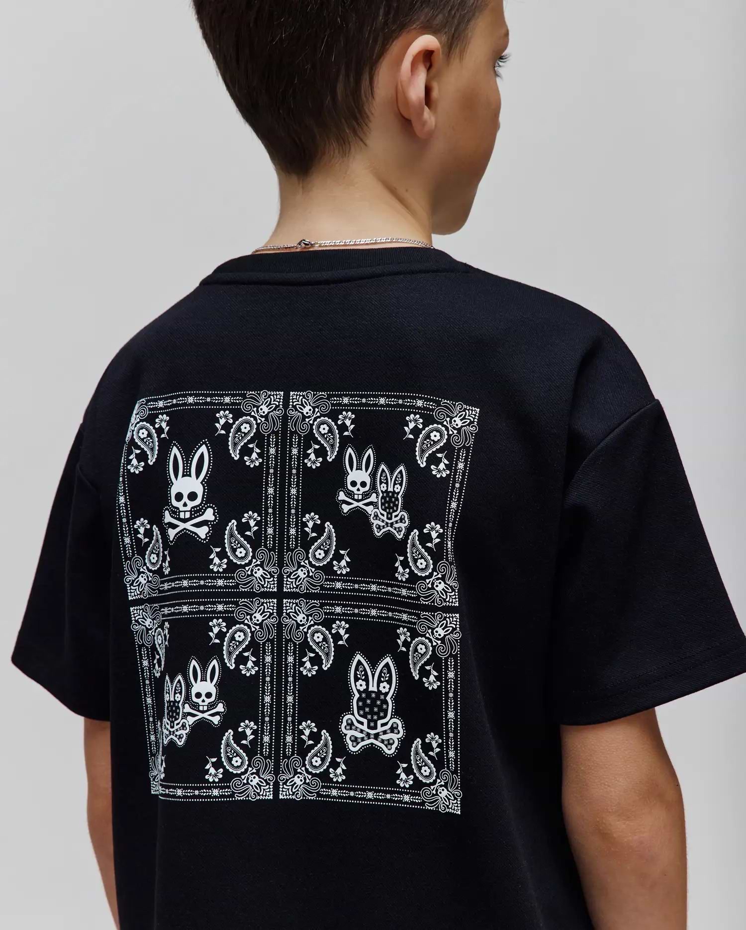 Kids Luke Back Graphic Tee, a black classic fit jersey t-shirt with a large white bandana-style graphic on the back featuring multiple Psycho Bunny skull logos and paisley patterns.