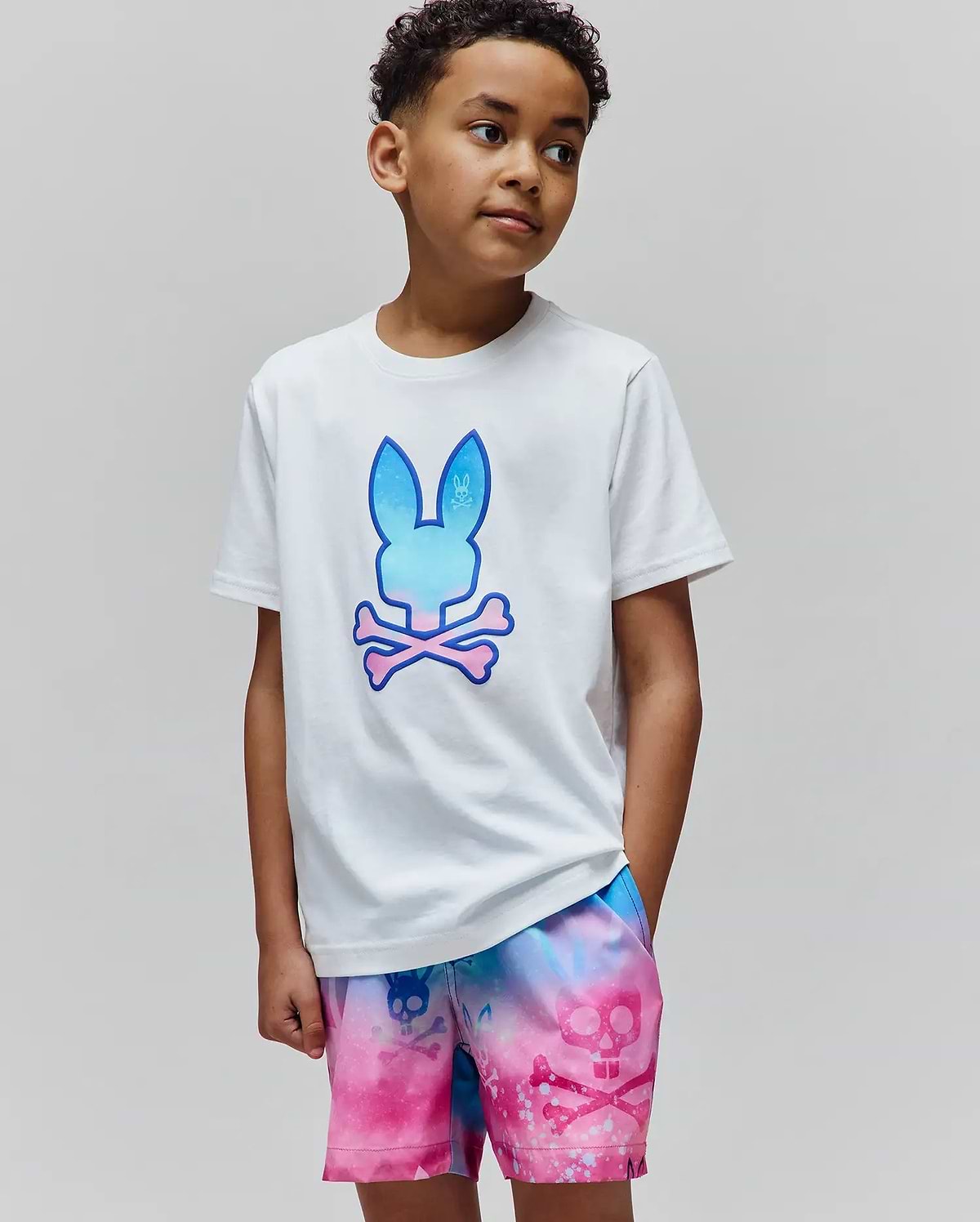 White Kids Caspian Graphic Tee, classic fit, soft jersey, with a large gradient blue-to-pink Psycho Bunny skull logo. Paired with vibrant tie-dye shorts featuring an all-over pink skull pattern.