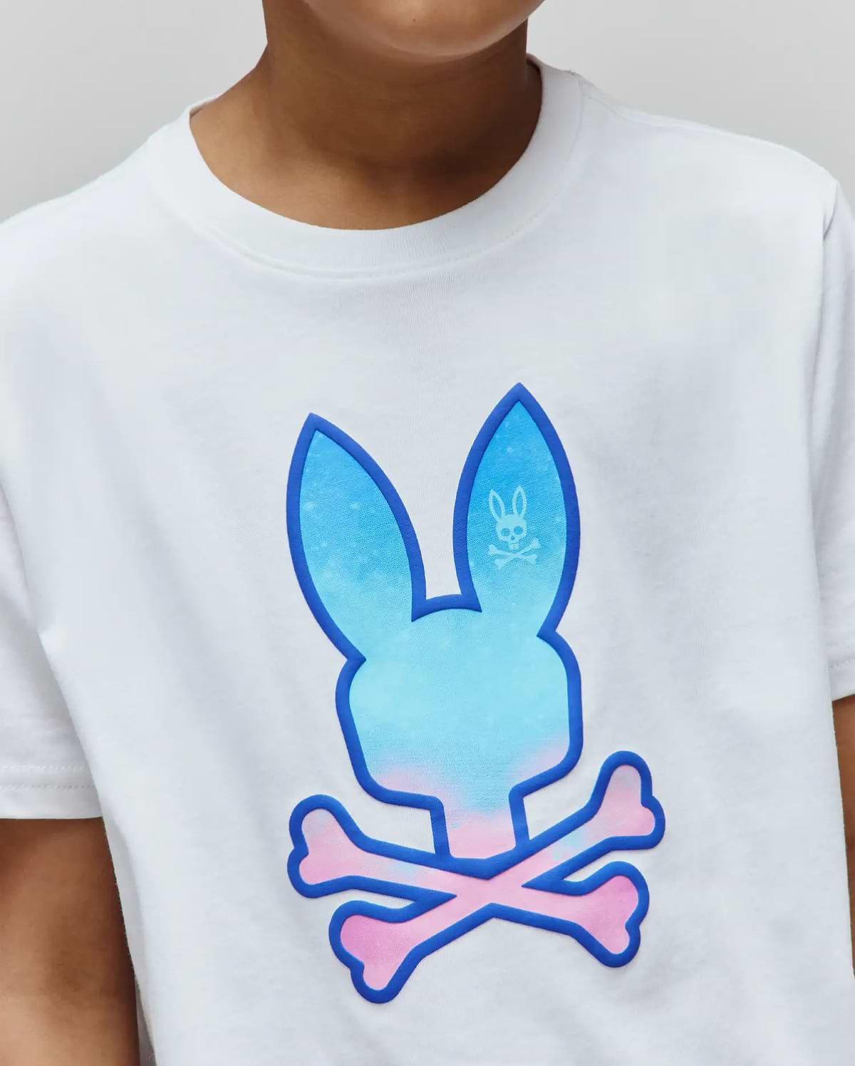 Kids Caspian Graphic Tee. White classic fit smooth jersey t-shirt with a large central Psycho Bunny logo featuring a blue to pink gradient and pink crossbones, outlined in dark blue.
