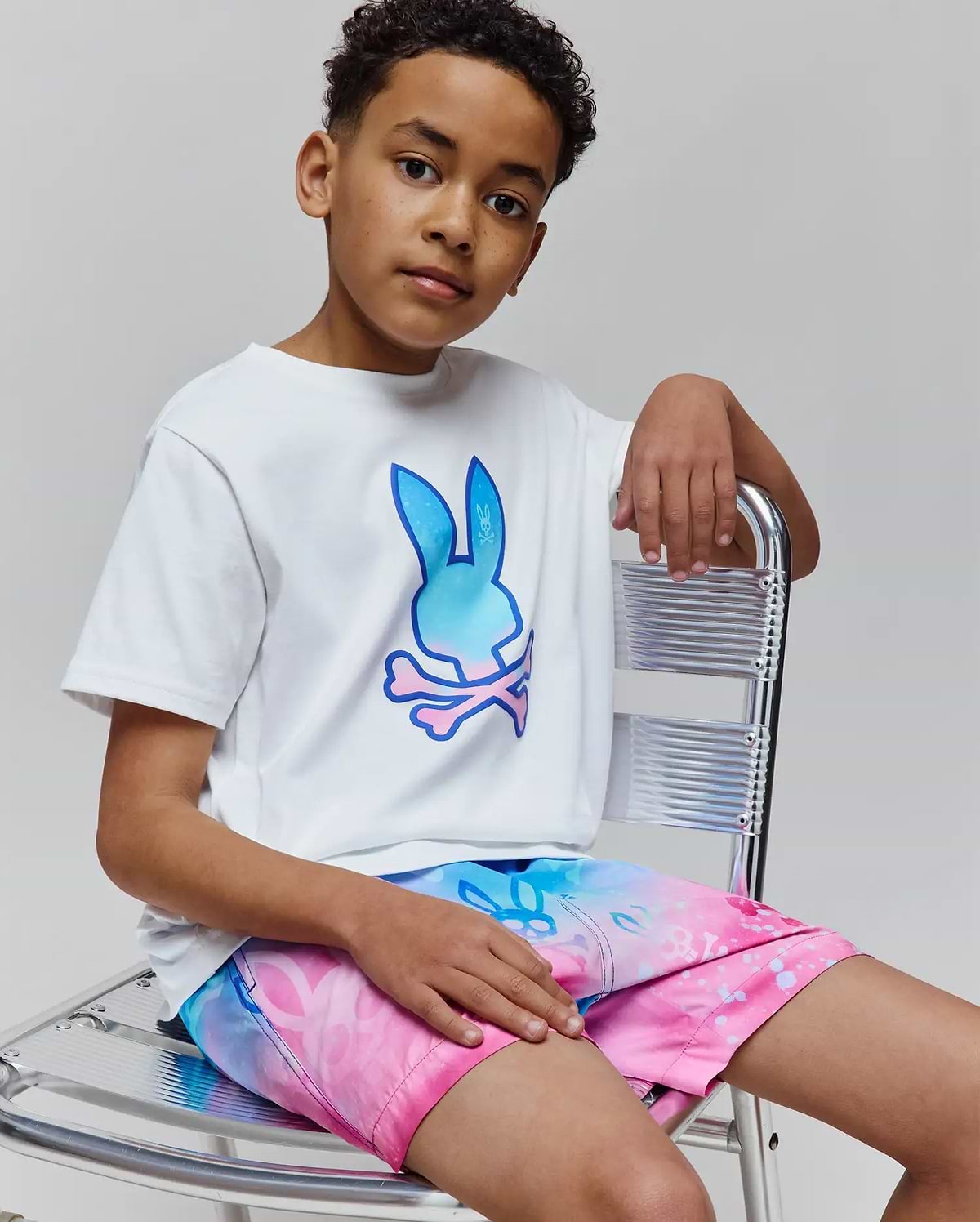 Kids Caspian Graphic Tee, a white classic-fit jersey t-shirt featuring a large gradient blue to pink Psycho Bunny skull logo on the chest, paired with vibrant gradient swim shorts.