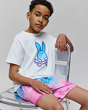 Kids Caspian Graphic Tee, a white classic-fit jersey t-shirt featuring a large gradient blue to pink Psycho Bunny skull logo on the chest, paired with vibrant gradient swim shorts.