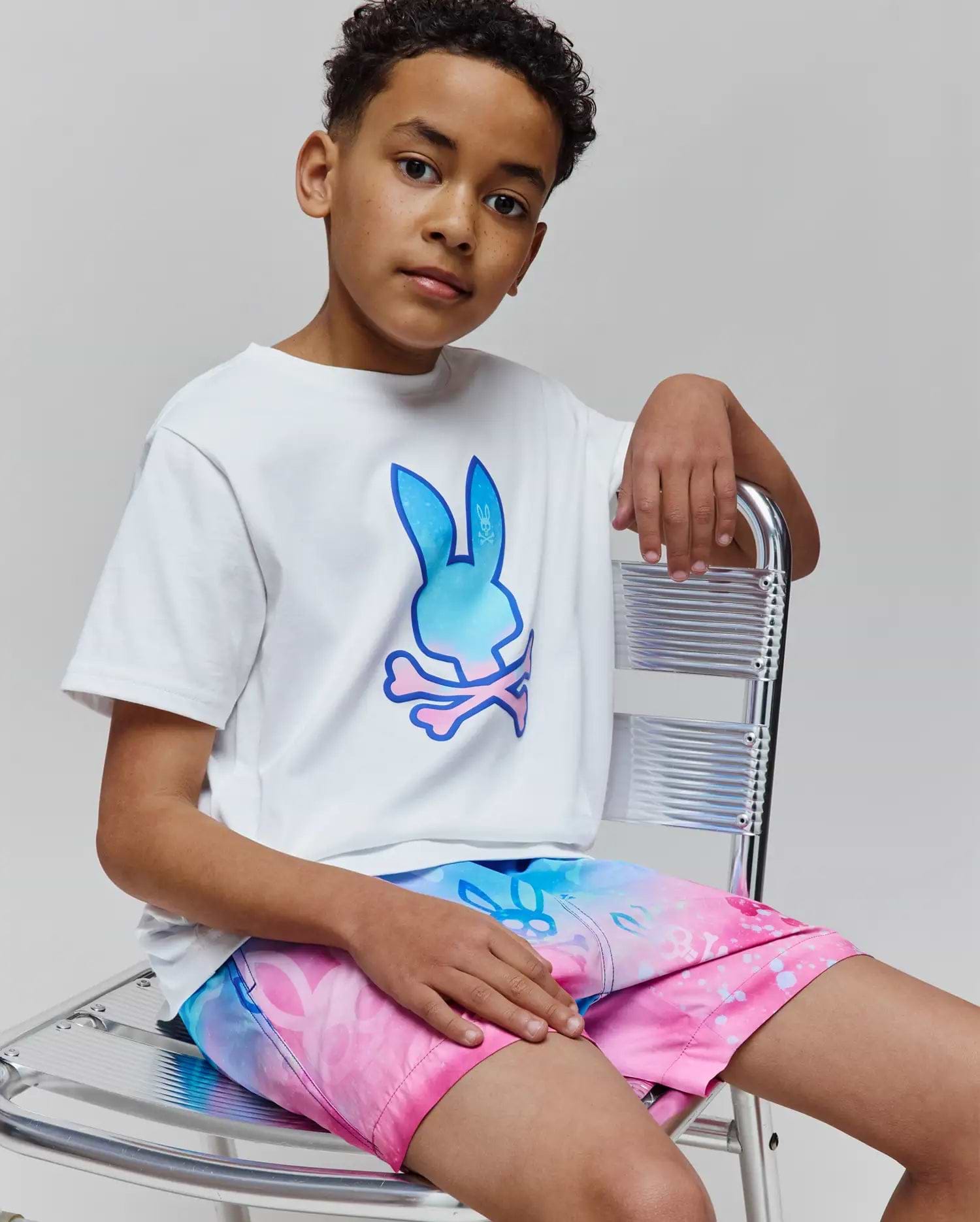 Kids Caspian Graphic Tee, a white classic-fit jersey t-shirt featuring a large gradient blue to pink Psycho Bunny skull logo on the chest, paired with vibrant gradient swim shorts.