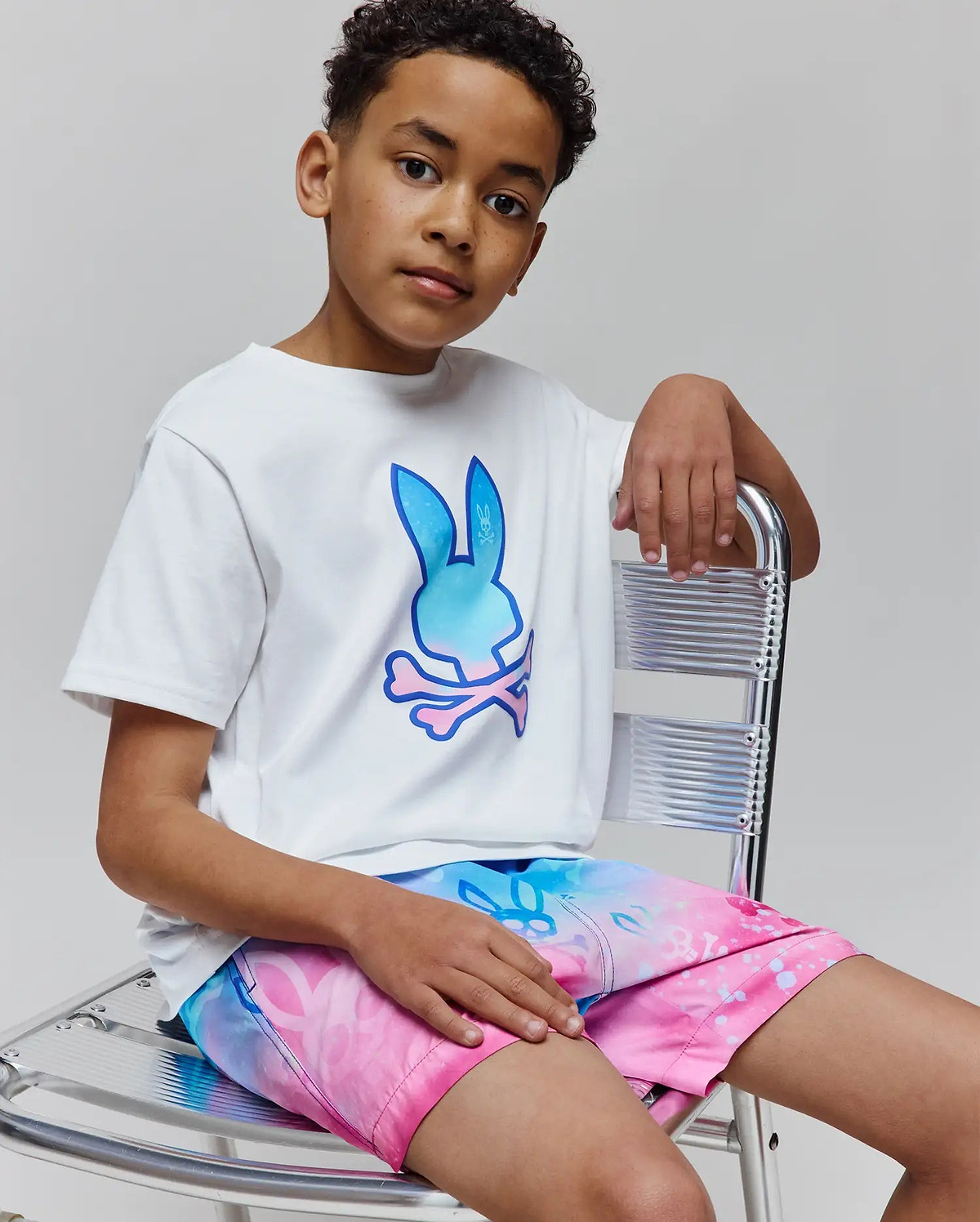 Kids Caspian Graphic Tee, a white classic-fit jersey t-shirt featuring a large gradient blue to pink Psycho Bunny skull logo on the chest, paired with vibrant gradient swim shorts.