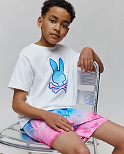Kids Caspian Graphic Tee, a white classic-fit jersey t-shirt featuring a large gradient blue to pink Psycho Bunny skull logo on the chest, paired with vibrant gradient swim shorts.