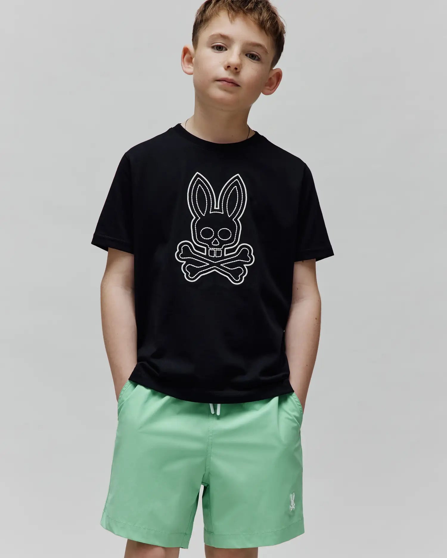 Black jersey Kids Marcel Graphic Tee with a classic fit, featuring a large white outline Psycho Bunny skull and crossbones graphic centered on the chest, paired with mint green shorts.
