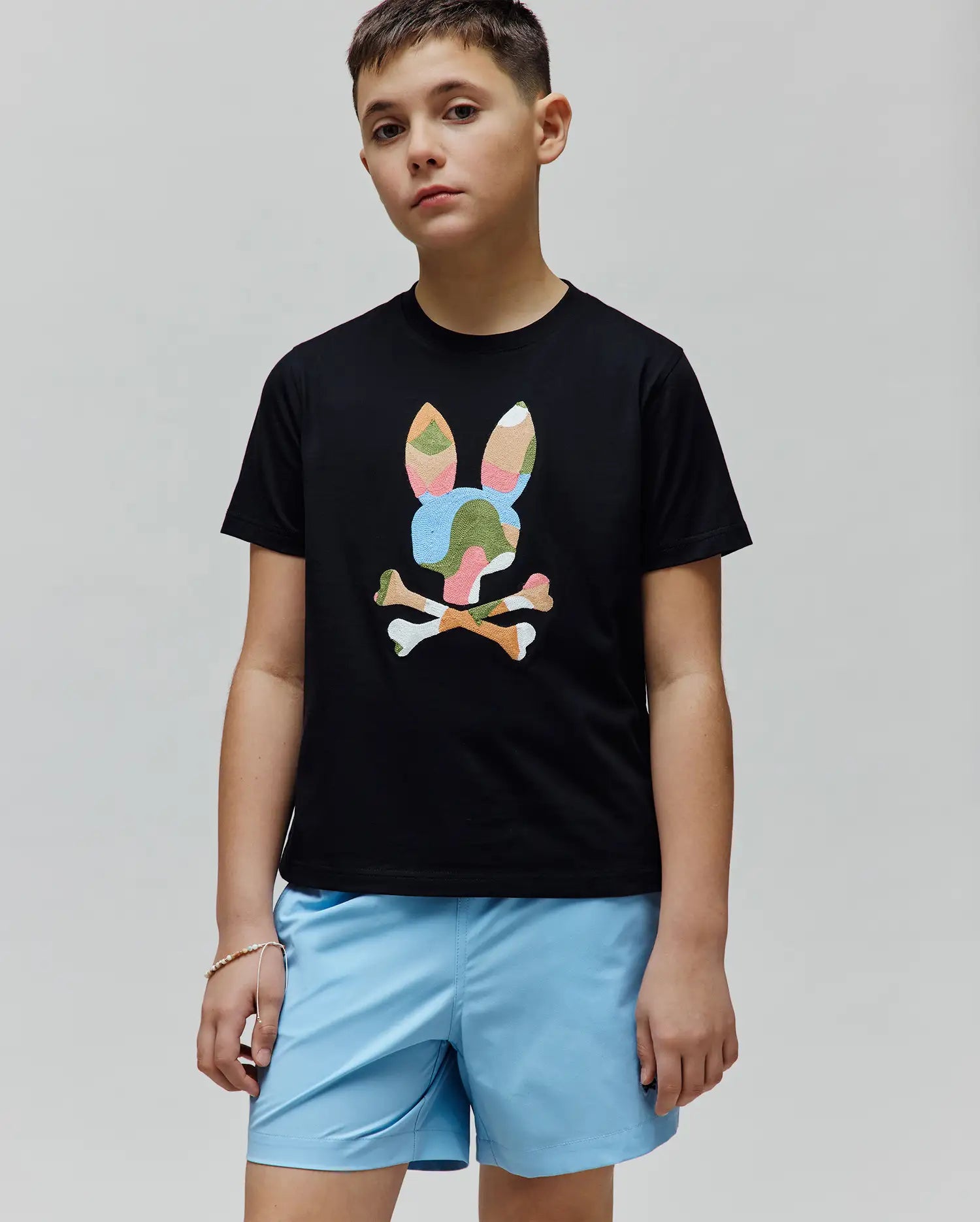 Kids Ara Graphic Tee, a black classic fit jersey t-shirt with a large central graphic of the iconic Psycho Bunny skull and crossbones logo in a vibrant multi-color camouflage pattern.