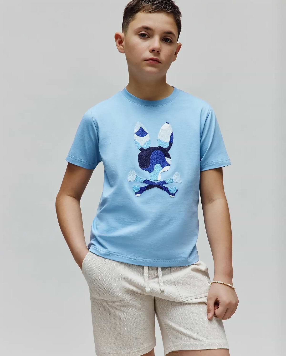 Kids Ara Graphic Tee in sky blue jersey knit, featuring a large central Psycho Bunny skull and crossbones logo with a multi-tonal blue camouflage pattern, classic fit.