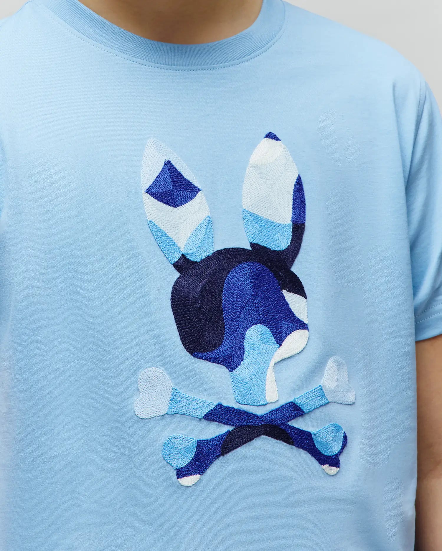 Kids Ara Graphic Tee, light blue classic fit jersey t-shirt with a large, central embroidered Psycho Bunny skull and crossbones logo, featuring a geometric blue and white patchwork design.