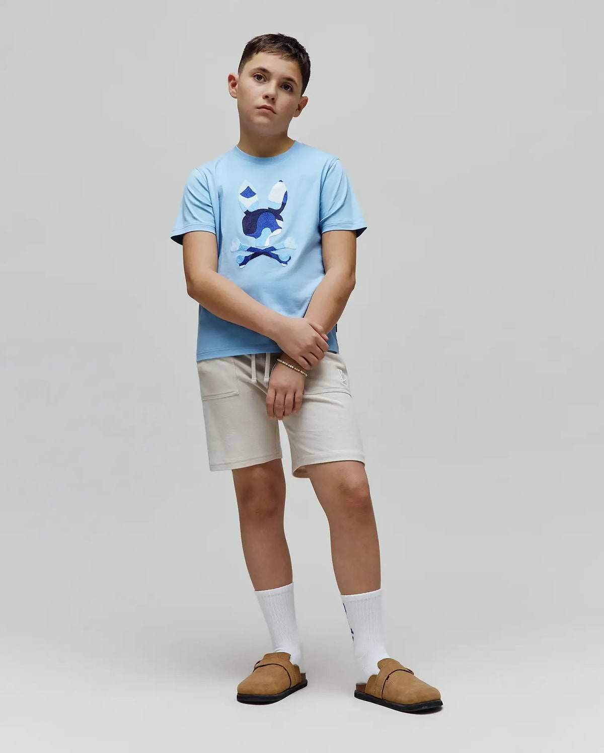 Kids Ara Graphic Tee in light blue jersey knit, featuring a classic fit and a large central chest graphic of the iconic Psycho Bunny skull and crossbones logo in multi-tone blue and white camouflage print.