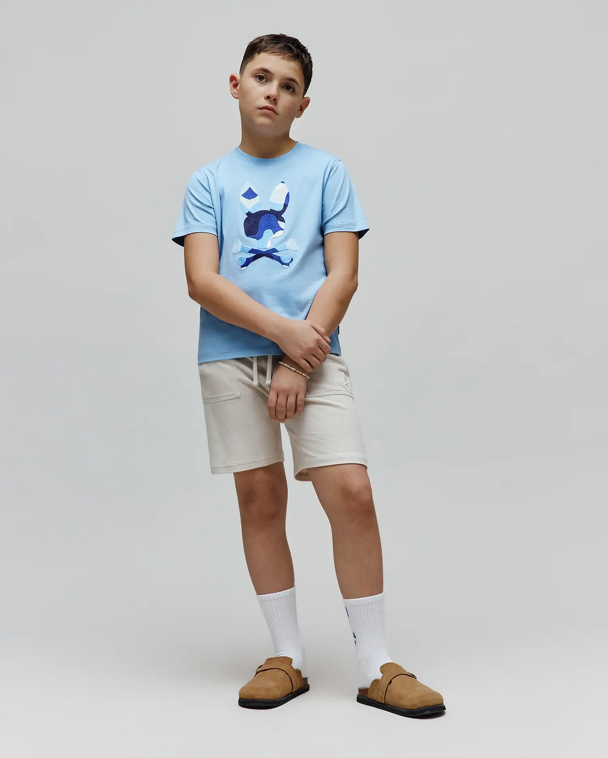 Kids Ara Graphic Tee in light blue jersey knit, featuring a classic fit and a large central chest graphic of the iconic Psycho Bunny skull and crossbones logo in multi-tone blue and white camouflage print.