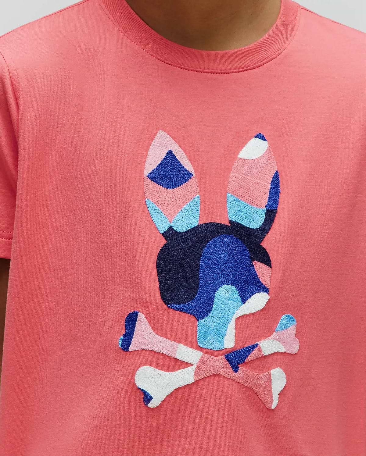 Kids Ara Graphic Tee, a coral classic fit jersey t-shirt with a large, central embroidered Psycho Bunny skull logo featuring a vibrant blue, light blue, pink, and white abstract pattern.
