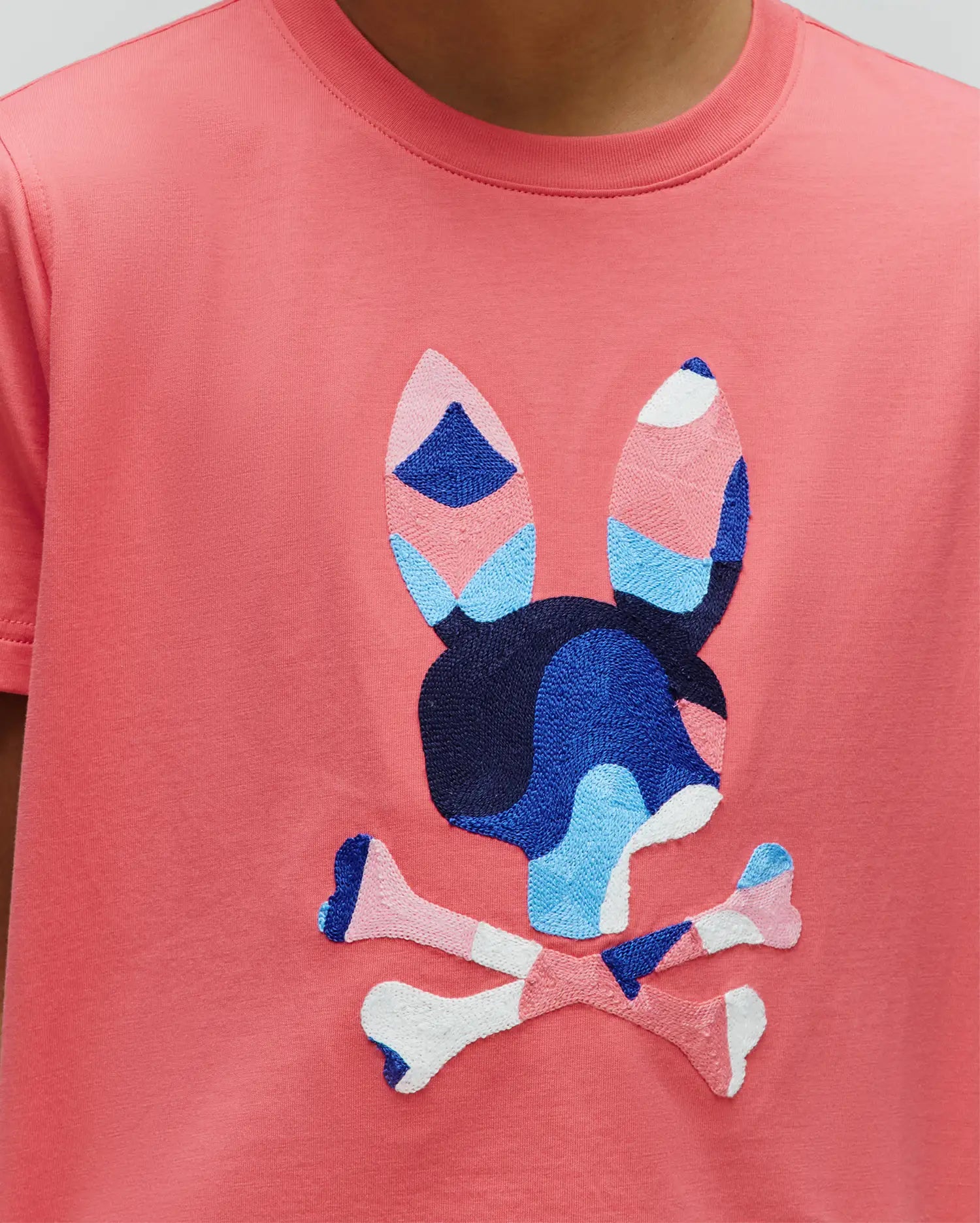 Kids Ara Graphic Tee, a coral classic fit jersey t-shirt with a large, central embroidered Psycho Bunny skull logo featuring a vibrant blue, light blue, pink, and white abstract pattern.
