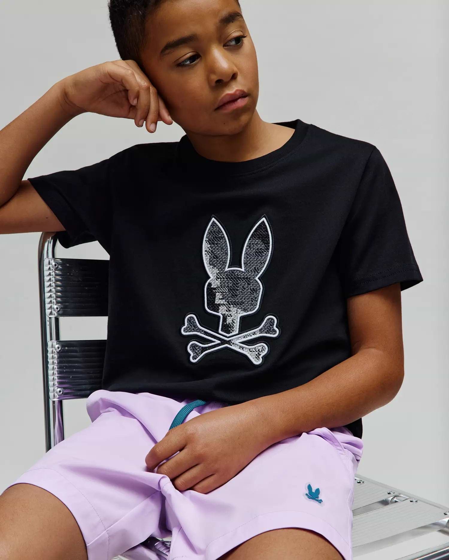 Kids Olivier Graphic Tee, a black classic fit jersey tee with a large sequined silver and white Psycho Bunny skull logo on the chest, paired with lavender woven shorts featuring a small teal embroidered bunny logo. (4)