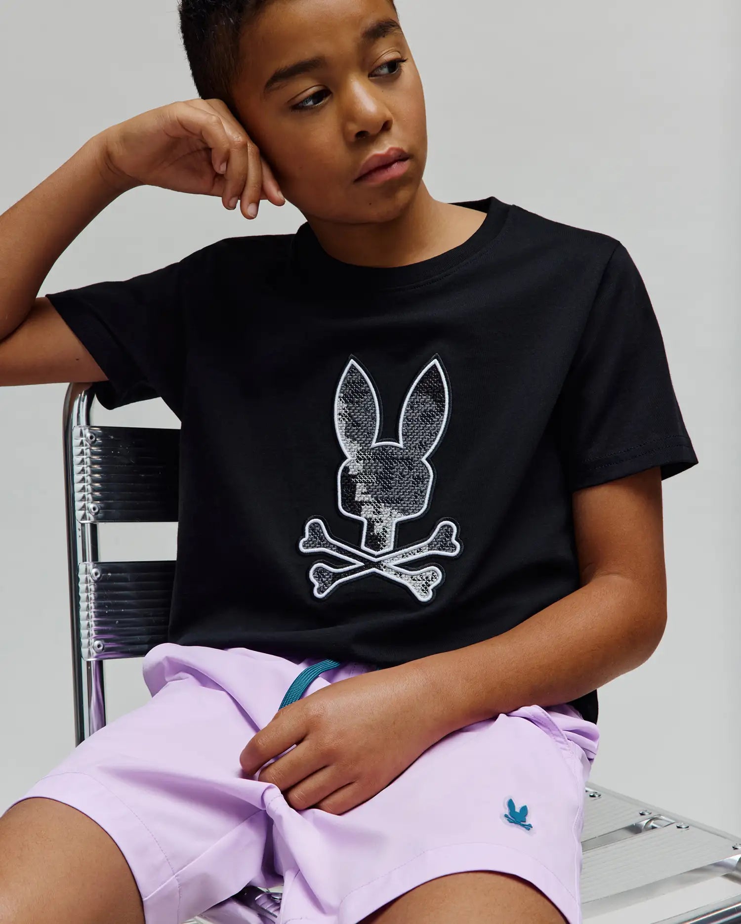 Kids Olivier Graphic Tee, a black classic fit jersey tee with a large sequined silver and white Psycho Bunny skull logo on the chest, paired with lavender woven shorts featuring a small teal embroidered bunny logo. (4)