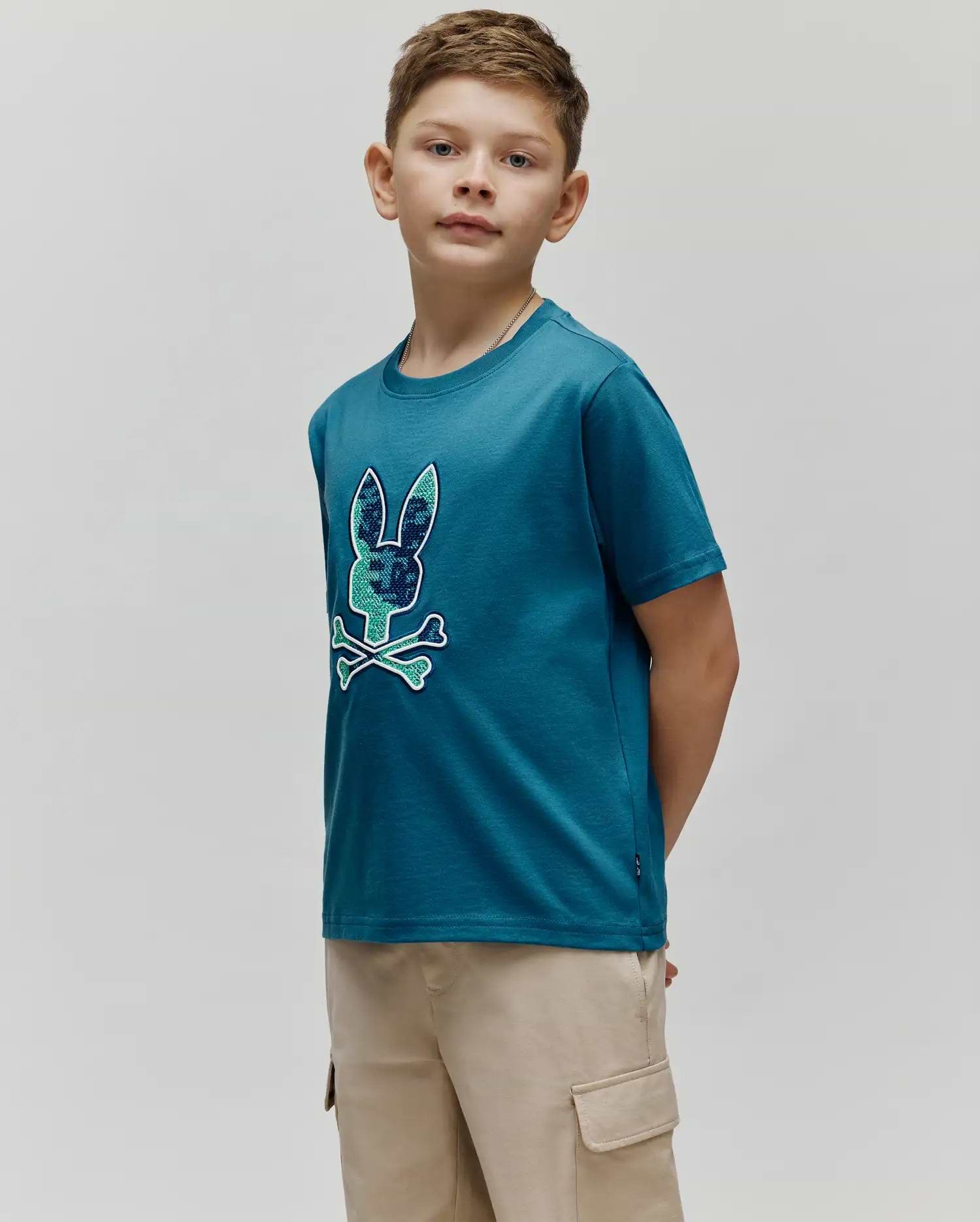 Kids Olivier Graphic Tee, a classic fit teal jersey t-shirt featuring a large chest graphic of the iconic Psycho Bunny skull logo with green and blue camouflage pattern and light blue bones, outlined in white. (2)