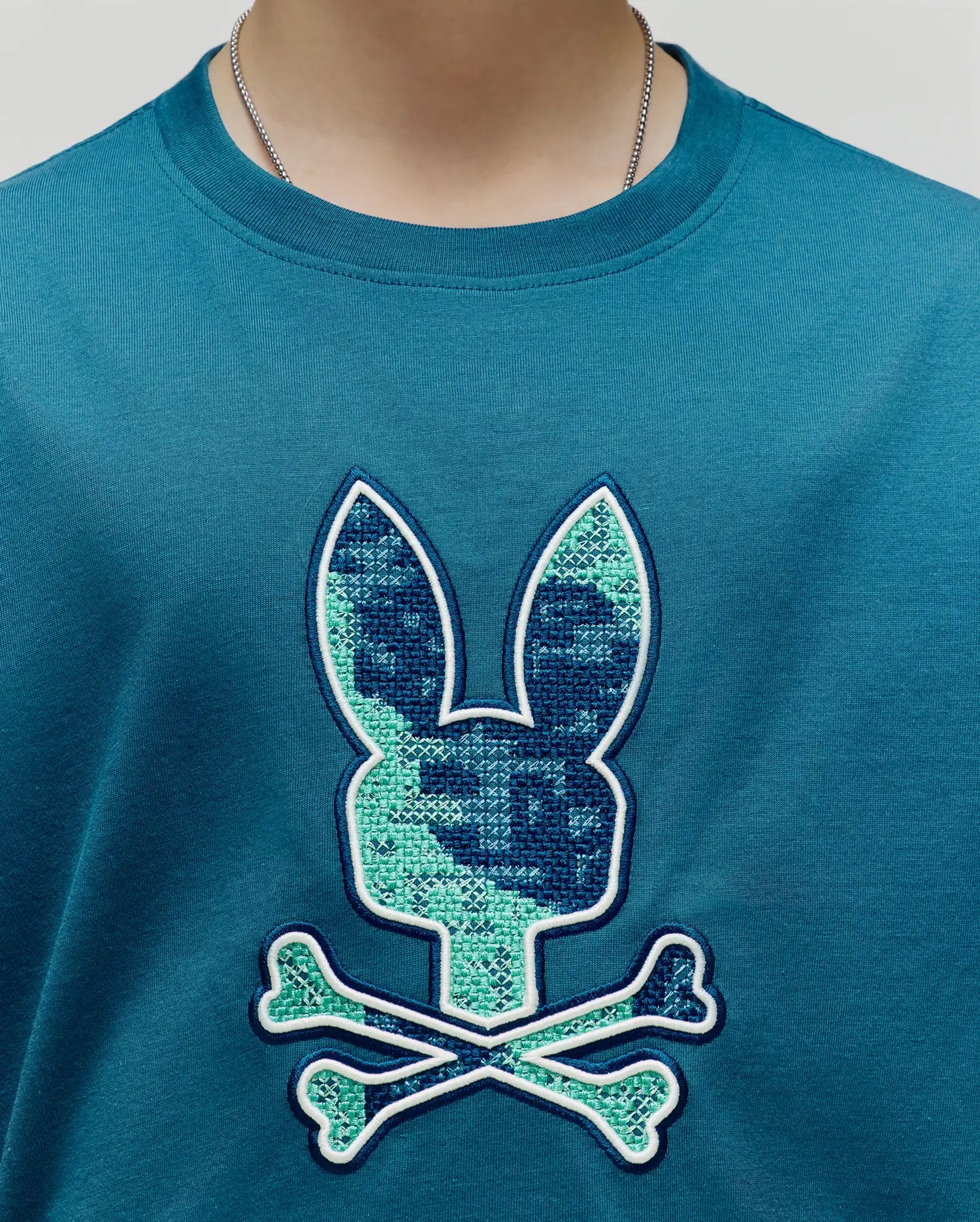 Kids Olivier Graphic Tee, a classic fit teal jersey knit t-shirt with a large central chest embroidered Psycho Bunny skull logo featuring a cross-stitch pattern in dark blue and light green, outlined in white. (3)