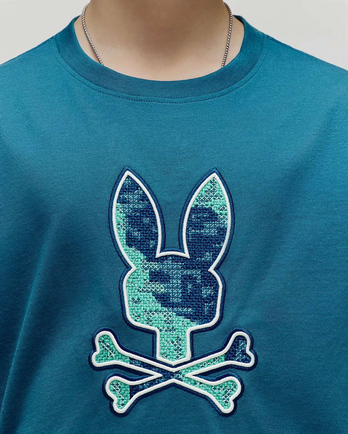 Kids Olivier Graphic Tee, a classic fit teal jersey knit t-shirt with a large central chest embroidered Psycho Bunny skull logo featuring a cross-stitch pattern in dark blue and light green, outlined in white. (3)