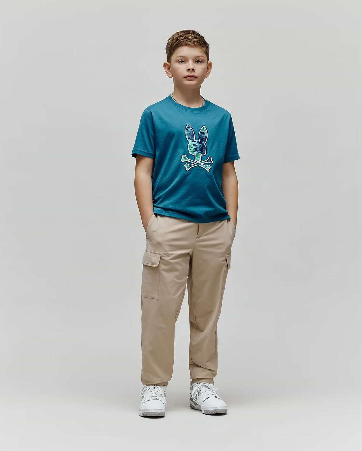 Kids Olivier Graphic Tee in teal jersey, featuring a large, textured blue and green Psycho Bunny skull and crossbones graphic on the chest, paired with relaxed fit khaki cargo pants. (4)