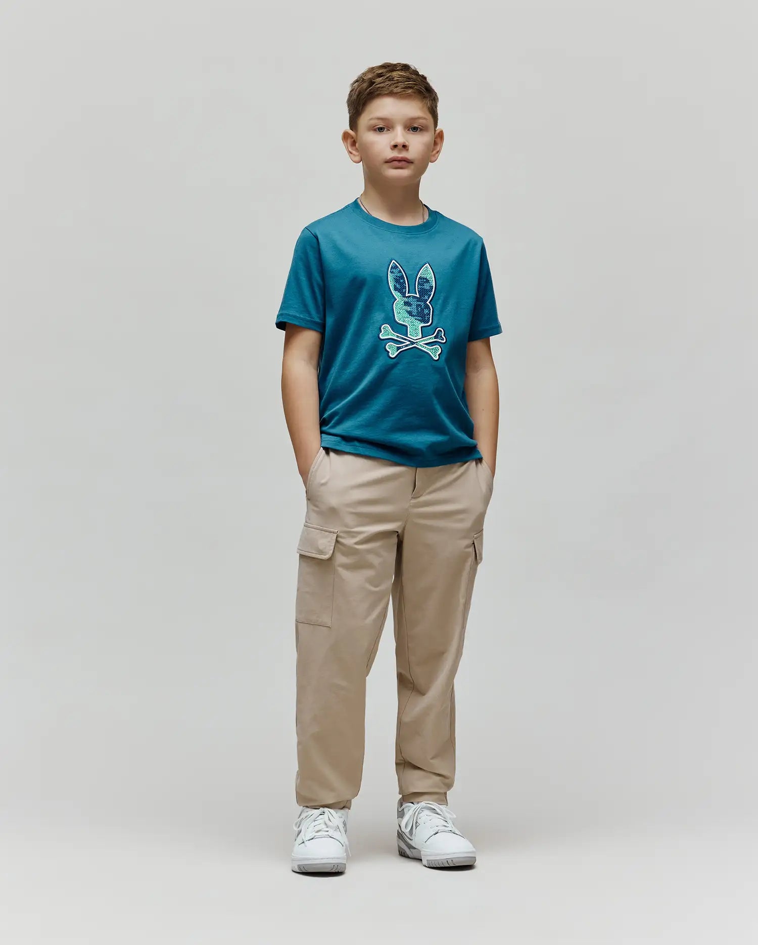 Kids Olivier Graphic Tee in teal jersey, featuring a large, textured blue and green Psycho Bunny skull and crossbones graphic on the chest, paired with relaxed fit khaki cargo pants. (4)
