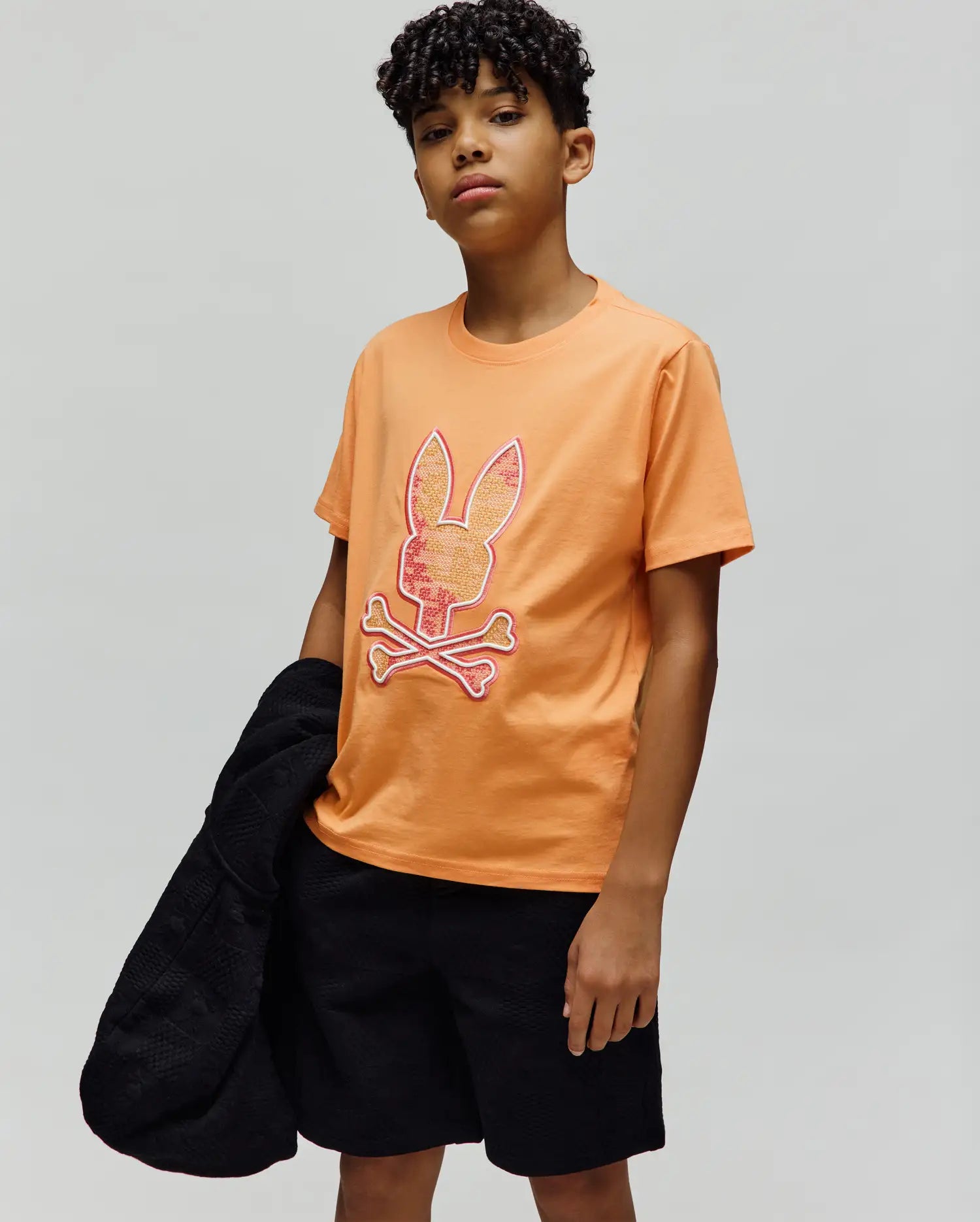 Kids Olivier Graphic Tee, a classic fit orange jersey t-shirt featuring a large central Psycho Bunny skull and crossbones logo with textured orange and red fill, outlined in red and white. (2)