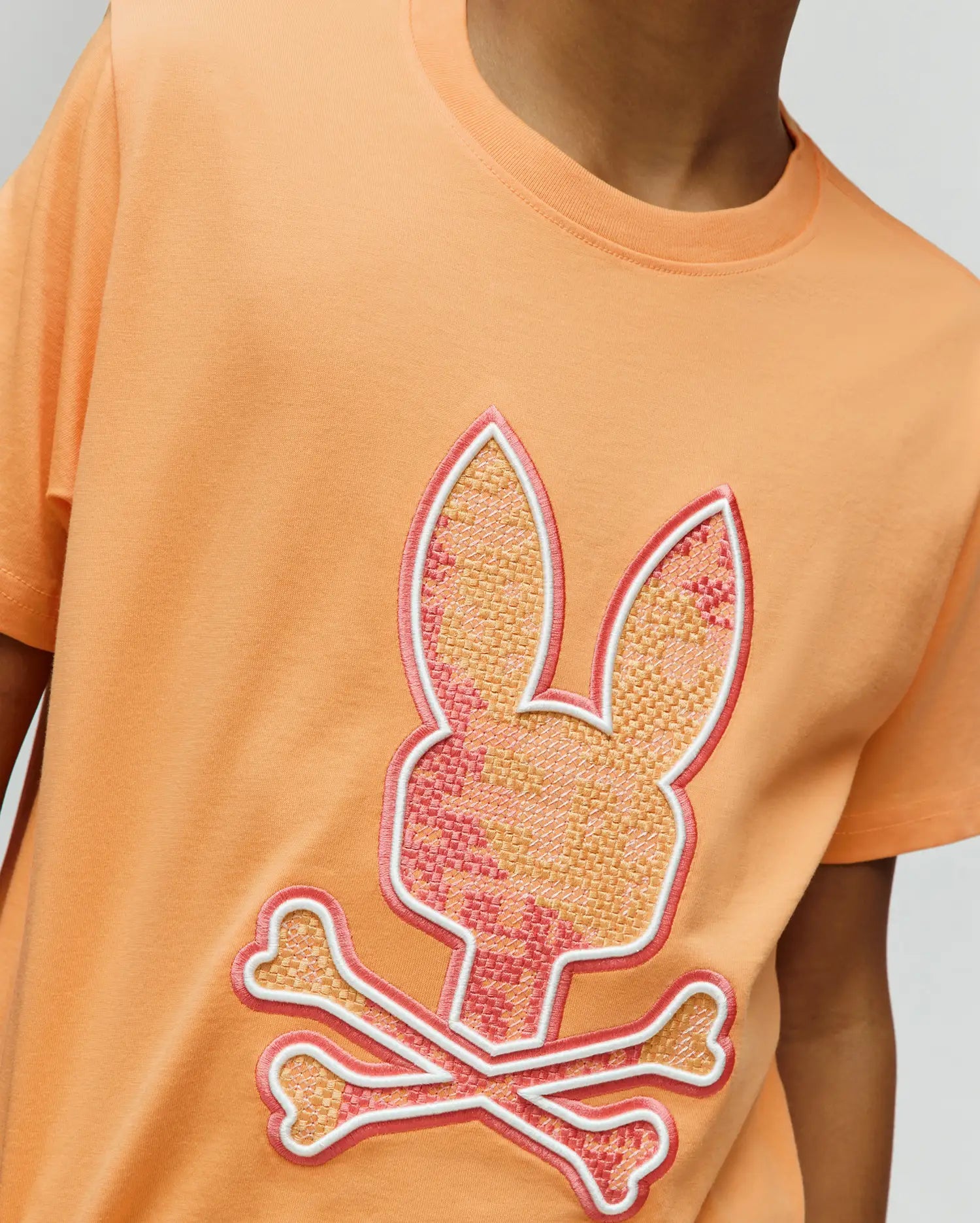 Kids Olivier Graphic Tee in peach, featuring a large, textured embroidered Psycho Bunny skull logo on the chest with a woven-like fill in pink and orange, outlined in white and pink, on a classic fit jersey knit. (3)