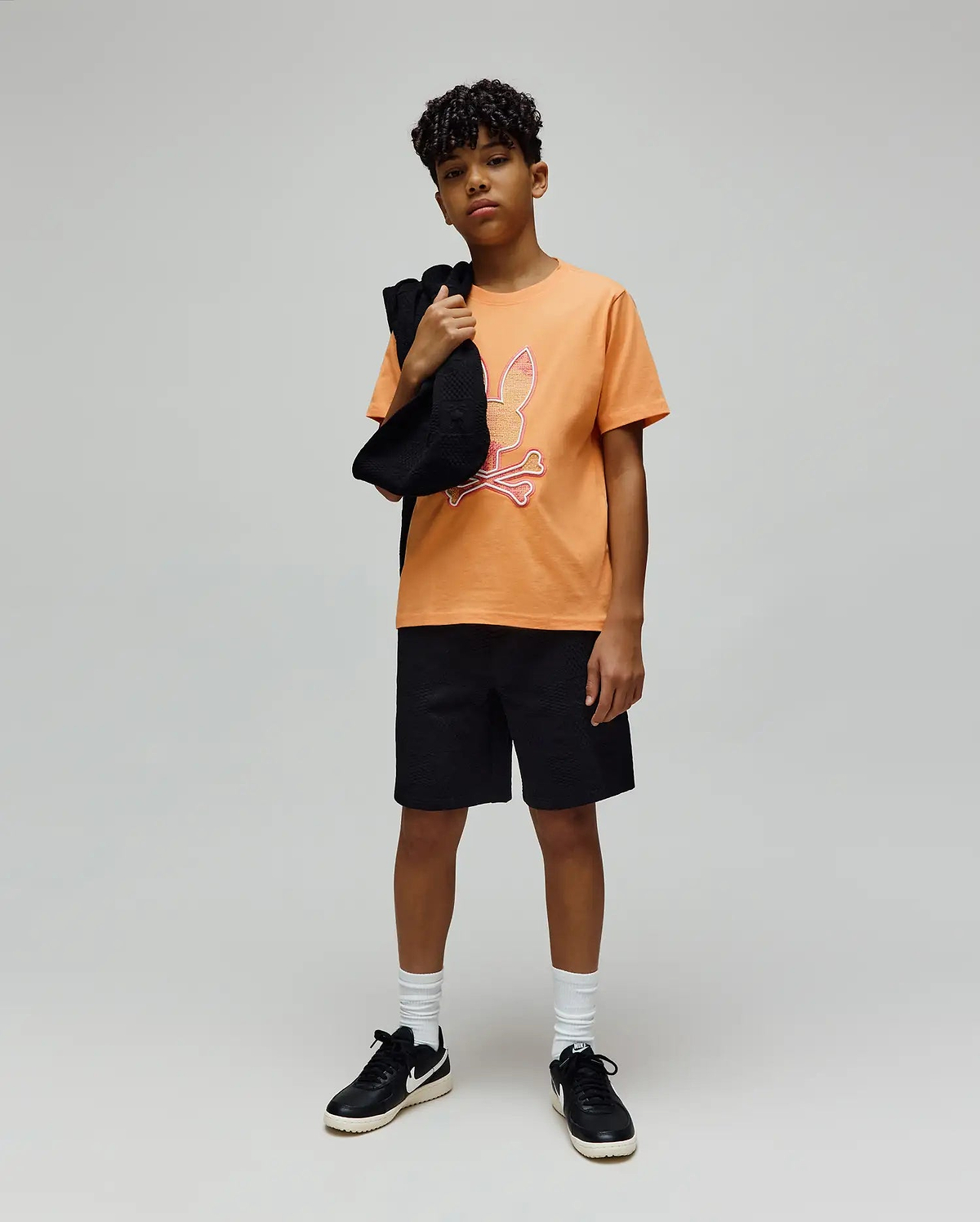 Kids Olivier Graphic Tee in vibrant orange jersey, featuring a large pink and white Psycho Bunny skull logo graphic on the chest, paired with black textured shorts for a relaxed fit. (4)