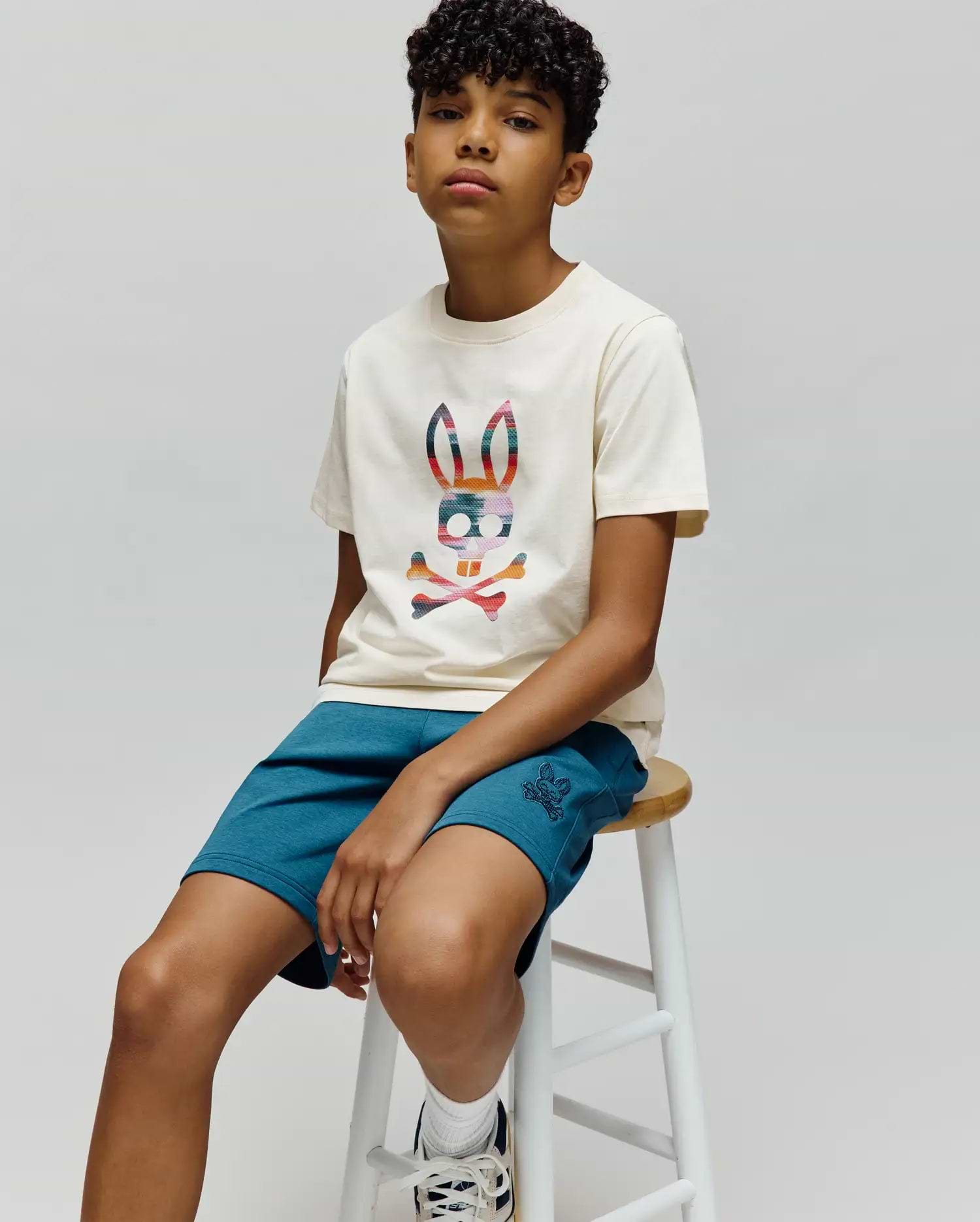 Kids Raphael Graphic Tee in cream, featuring a large colorful plaid Psycho Bunny skull graphic on the chest, paired with teal jersey shorts with a small embroidered bunny logo.