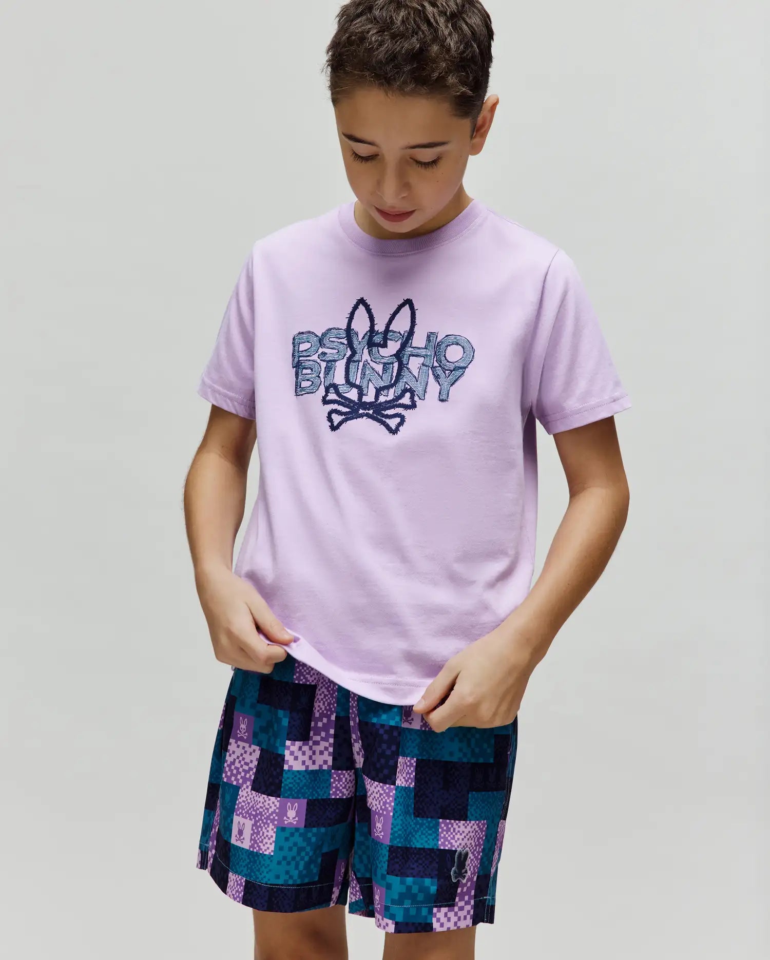 Kids Herman Graphic Tee in smooth lavender jersey, featuring a large pixelated blue Psycho Bunny logo on the chest, paired with vibrant multi-color pixelated shorts with subtle bunny skull branding. (2)