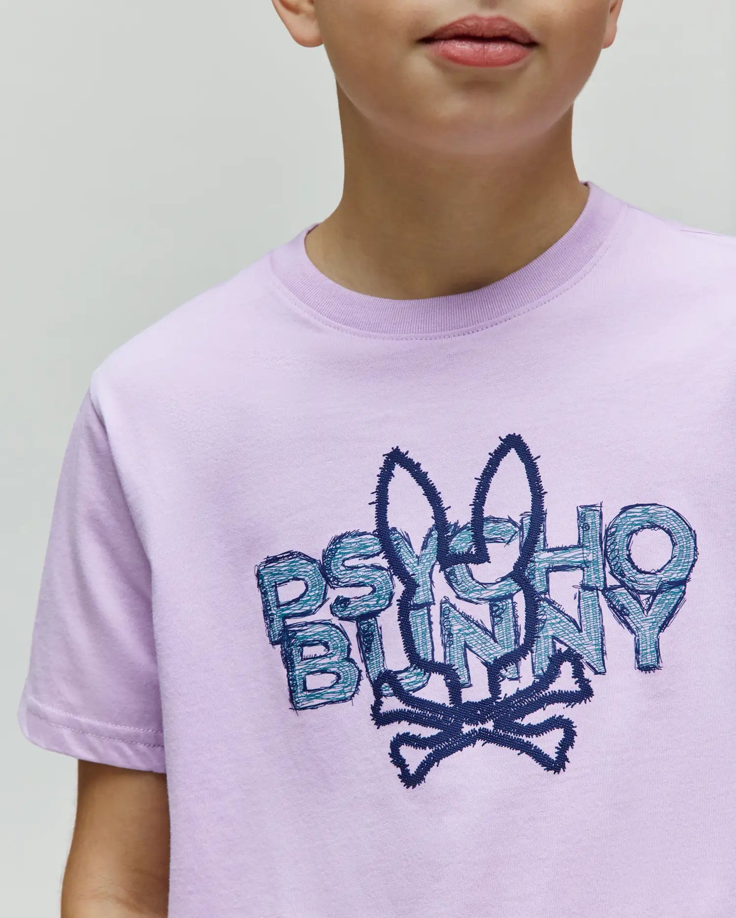 Kids Herman Graphic Tee, a light purple jersey t-shirt with a large, central embroidered Psycho Bunny skull and crossbones logo in dark blue and teal, featuring textured lettering. (3)