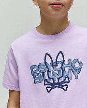 Kids Herman Graphic Tee, a light purple jersey t-shirt with a large, central embroidered Psycho Bunny skull and crossbones logo in dark blue and teal, featuring textured lettering. (3)