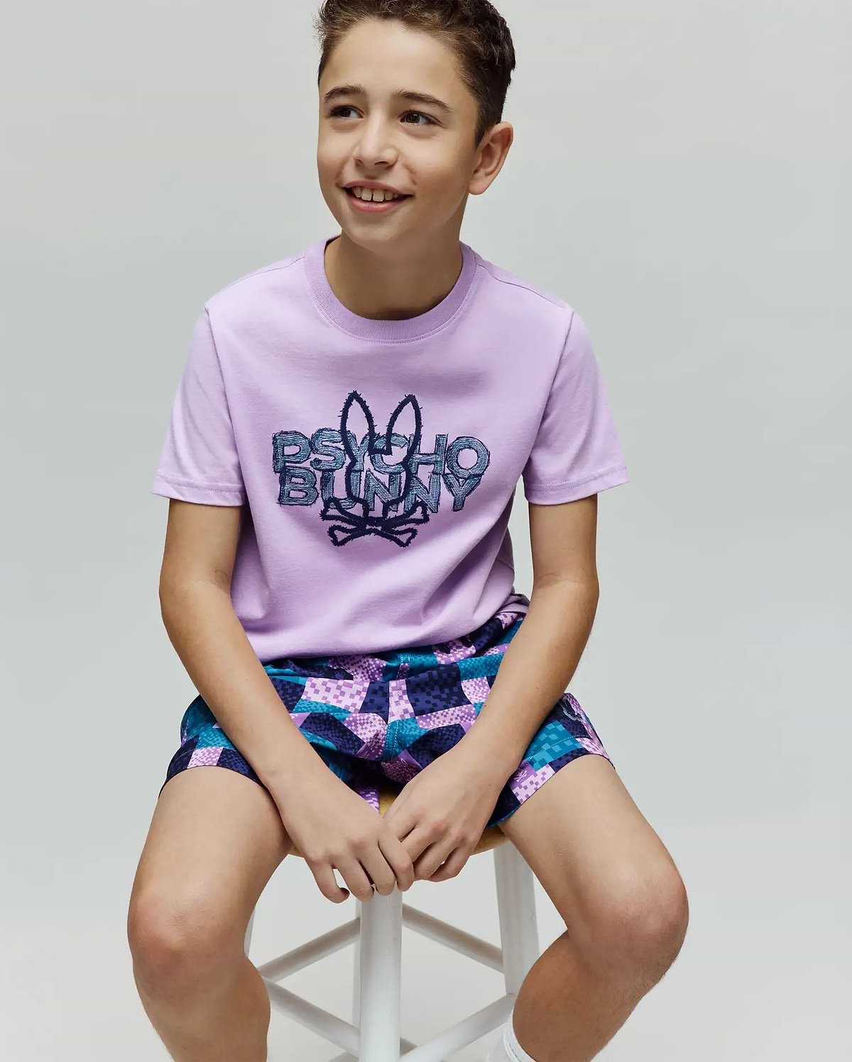Kids Herman Graphic Tee, a classic fit lilac jersey t-shirt featuring a large central textured Psycho Bunny skull logo and text graphic, paired with relaxed geometric patchwork shorts in purple, teal, and blue. (4)